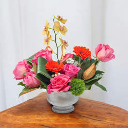 Pink roses and orange gerberas in a gray vase with a tall orchid stem