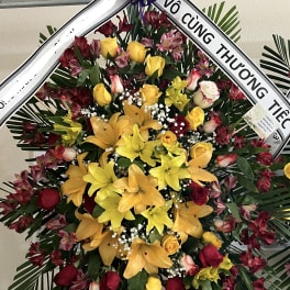 Large standing floral wreath with yellow lilies, red roses, and a purple ribbon