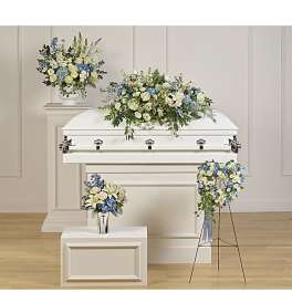 White casket surrounded by blue and white floral arrangements, including a casket spray and heart wreath on a stand.