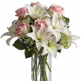 Tall glass vase with white lilies and light pink roses arranged with small white filler flowers.