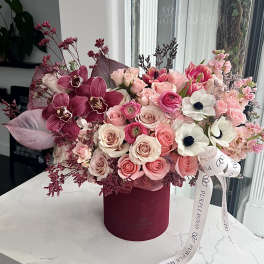Pink and burgundy floral arrangement with roses, orchids, and white anemones in a round velvet hatbox.