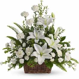 White lilies and carnations arranged with ferns in a brown wicker basket.