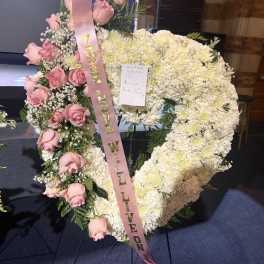 Heart-shaped floral tribute with pink roses and white flowers, tied with a pink ribbon