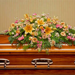 Low spray of yellow lilies and mixed pink flowers arranged atop a wooden casket