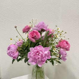 Pink peonies and roses with small white daisies arranged in a ribbed glass vase on a white pedestal