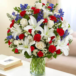 Tall bouquet of white lilies, white roses, red carnations, and blue flowers in a clear glass vase