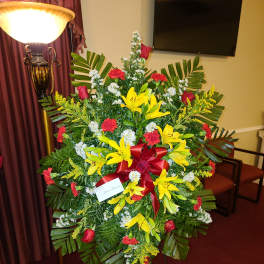 Large standing floral spray with yellow lilies, red roses, and a red ribbon