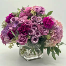 Low arrangement of pink and purple roses, carnations, mums, and hydrangeas in a clear glass cube vase