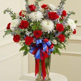 Patriotic red, white, and blue bouquet with roses, carnations, and mums in a clear glass vase with ribbon bow