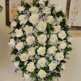 Oval standing spray of white roses and baby's breath on an easel with a black dedication sign.
