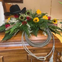 Cowboy hat atop a floral arrangement with roses and sunflowers on a wooden casket.