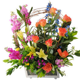 Tall mixed arrangement of orange roses, yellow lilies, and bright blooms in a white rectangular container