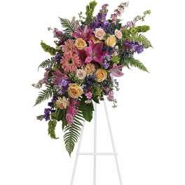 Asymmetrical standing spray of pink lilies, gerbera daisies, and roses with purple blooms on a white easel.