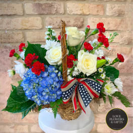 Patriotic basket arrangement with white roses, red carnations, blue hydrangea, and daisies with flag ribbon