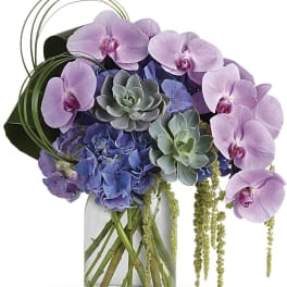 Lavender orchids and blue hydrangeas in a clear glass vase with succulents