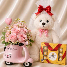 Pink rose bouquet in a scooter-shaped vase beside a teddy bear and chocolates