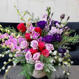 Low bowl arrangement of pink and red roses with purple blooms and orchids in a lush mixed design