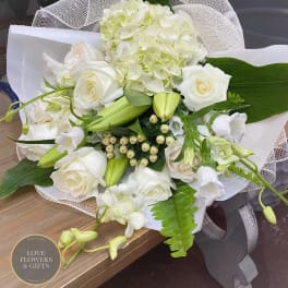 Hand-tied bouquet of white roses, lilies, orchids, and hydrangea wrapped in white mesh and paper