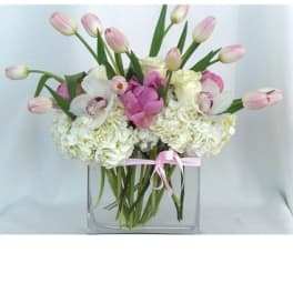 Pink tulips and white flowers arranged in a clear square vase with a pink ribbon