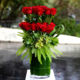 Red roses arranged in a tall glass vase with succulents and fern-like foliage