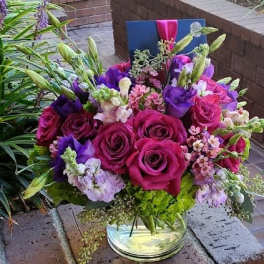 Low glass vase of fuchsia roses, purple blooms, and mixed pink flowers with a card tucked at the back.