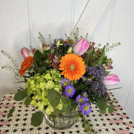 Mixed bouquet with orange gerbera daisies, pink tulips, and purple flowers in a glass vase