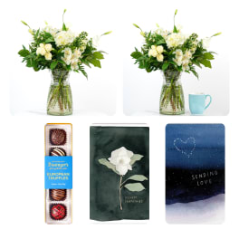 White floral arrangement in a glass vase with a chocolate box and sympathy cards nearby