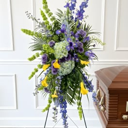 Tall funeral spray with blue, purple, green, and yellow flowers beside a casket