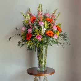 Tall arrangement of pink roses, orange gerbera daisies, lilies and snapdragons in a clear glass vase