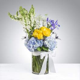 Bouquet of yellow roses, blue hydrangeas, and purple irises in a glass vase with a white ribbon.