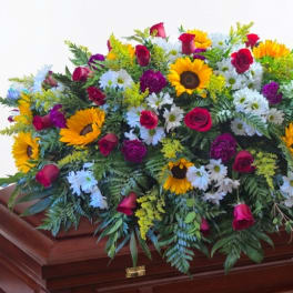 Large floral spray with sunflowers, roses, and white daisies on a wooden casket
