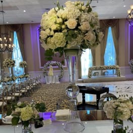 Tall white rose and hydrangea centerpiece in a clear glass vase on a mirrored table.