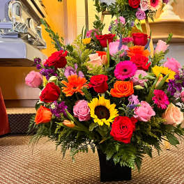 Tall mixed arrangement of bright roses, gerbera daisies, and sunflowers in a black vase