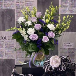 Lavender roses and white flowers arranged in a glass vase with ribbons.