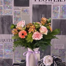 Pink and peach mixed flowers arranged in a white vase with a lavender ribbon.