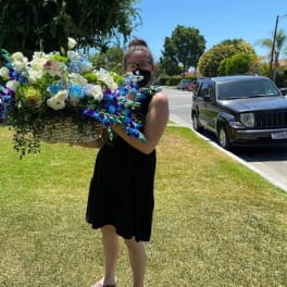 Person in a black dress holding a large basket of white, blue, and purple flowers on a lawn