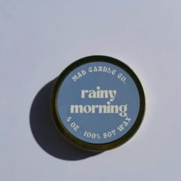 Round candle tin labeled "rainy morning" on a plain surface