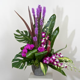 Tropical arrangement with purple orchids and tall purple spikes in a gray vase
