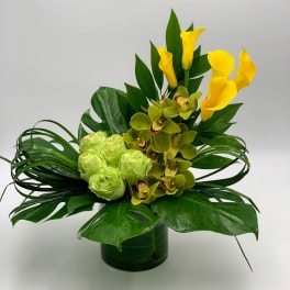 Yellow calla lilies and green orchids in a glass vase with broad leaves