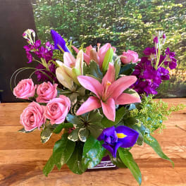 Pink lilies and roses arranged with purple flowers in a low container