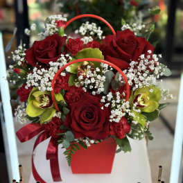 Red roses and orchids arranged in a red handled box with a ribbon