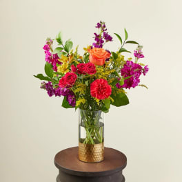 Tall mixed bouquet of bright pink, orange, and yellow flowers in a glass vase with gold base
