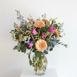 Mixed bouquet of peach gerberas, purple tulips, and blue flowers in a clear glass vase.