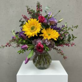 Bouquet of yellow gerbera daisies, pink roses, and purple flowers in a glass vase