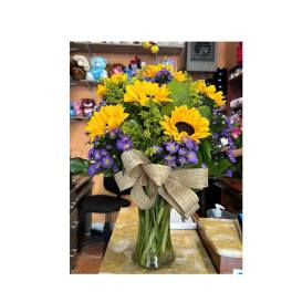 Sunflower and purple daisy bouquet in a glass vase with a burlap bow