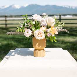Low arrangement of white and peach roses in a matte yellow pedestal vase set outdoors.