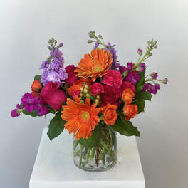 Mixed bouquet of orange gerbera daisies, pink roses, and purple flowers in a glass vase