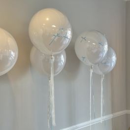 Clear balloons with pale blue ribbons and long white streamers against a wall