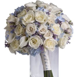 Round bridal bouquet of white roses, calla lilies, and soft blue accents displayed in a clear glass vase