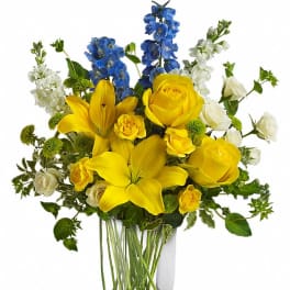 Tall glass vase of yellow lilies and roses with blue delphinium and white blooms